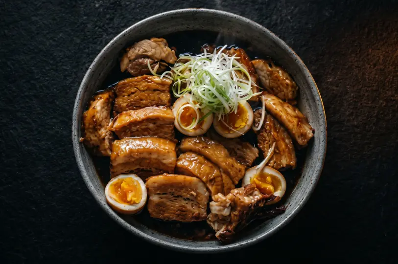 Top-down view of a bowl filled with thick slices of braised pork belly (kakuni) and halved soft-boiled eggs resting in a rich, dark broth, garnished with freshly sliced scallions.