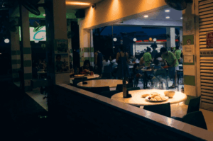 An atmospheric night scene at a bustling open-air eatery. Warm light illuminates a round, white table in the foreground holding a plate of sliced meat and a bowl of soup. In the background, patrons dine and socialize at tables under the glow of overhead lights against the dark evening sky.