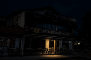 A dark, moody nighttime photograph of a two-story corner building in Singapore. The ground floor features the sign for R.K. Eating House, while the second floor houses the Bulldog Cafe-Pub. Most of the scene is draped in deep shadow, except for a solitary pool of warm, golden light illuminating a cluster of empty outdoor dining tables and chairs, capturing the quiet stillness of the city late at night.