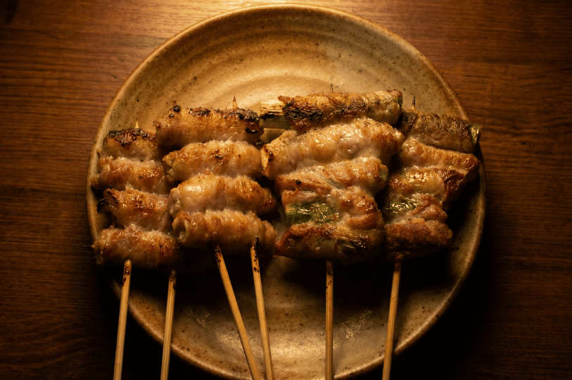 A close-up of five grilled skewers featuring thin slices of meat wrapped around vegetables, served on a rustic ceramic plate under warm, moody lighting typical of an izakaya setting.