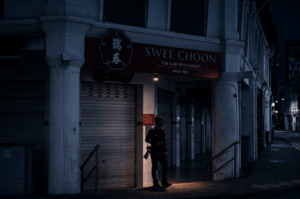 A moody, late-night scene featuring a lone photographer standing outside the closed roller shutters of the iconic Swee Choon Tim Sum Restaurant. The figure, holding a camera, is illuminated by a single warm downlight that cuts through the cool, dark blue shadows of the empty five-foot way and historic shophouse architecture, capturing the quiet solitude and anticipation of urban exploration after dark.