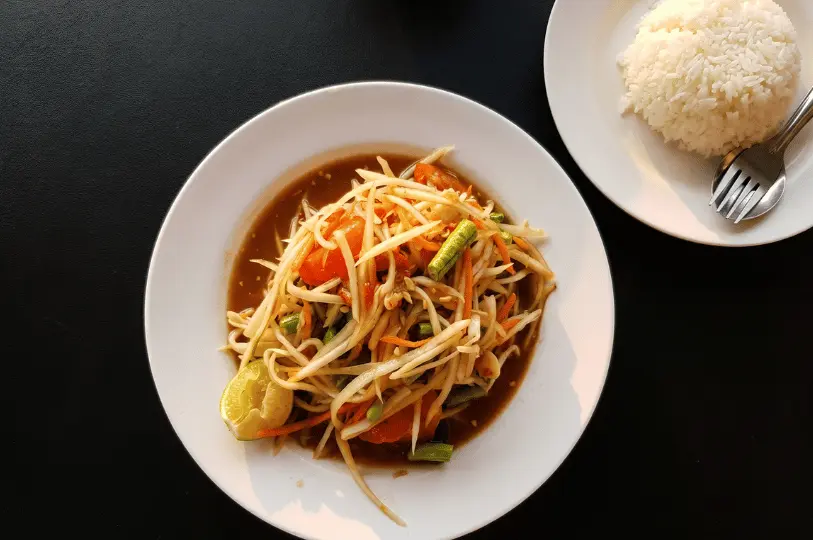 A top-down view of a vibrant plate of Som Tum (Thai green papaya salad) with tomatoes and green beans, served alongside a separate plate of white rice.