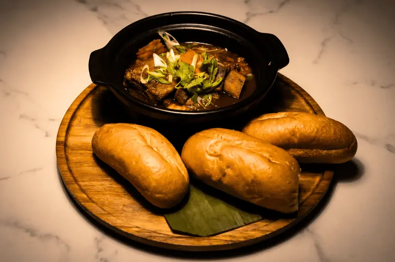 A rustic meal served on a round wooden board featuring a dark claypot filled with hearty braised meat and carrot stew topped with fresh herbs, accompanied by three plain, crusty bread rolls.