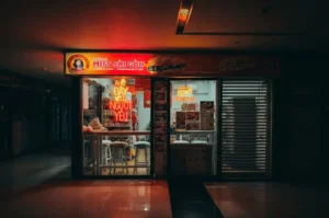 The brightly lit storefront of the Miss Saigon eatery in a darkened, quiet mall corridor. Glowing red and orange neon signs illuminate a small dining counter with empty stools, capturing a late-night atmosphere.