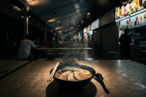 A steaming bowl of ramen sits in a dramatic pool of light on a textured tabletop, standing out sharply against the deep shadows and moody, dimly lit background of a quiet, late-night open-air food court.