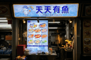 A brightly illuminated hawker stall named "Tian Tian You Yu" operating at night. The stall's glowing blue and white signboard features a cartoon cat holding a fishing rod alongside large Chinese characters. Below the sign, a tall vertical light-up menu displays photos of various colorful seafood and fish soup dishes. The front counter holds stacks of red baskets, utensils, and condiment containers, while the background reveals a glimpse into the working stainless-steel kitchen area, capturing the vibrant, practical energy of late-night dining in Singapore.