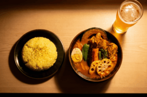 A top-down view of a hearty bowl of Hokkaido soup curry filled with chicken, sliced lotus root, pumpkin, green pepper, and a boiled egg half. It sits on a wooden table next to a black plate of seasoned yellow rice and a glass of frothy beer, all illuminated by warm, dramatic overhead lighting.