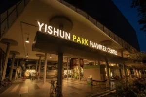 Exterior shot of Yishun Park Hawker Centre at night, showcasing the large illuminated sign at the entrance with outdoor seating beneath warm lighting.