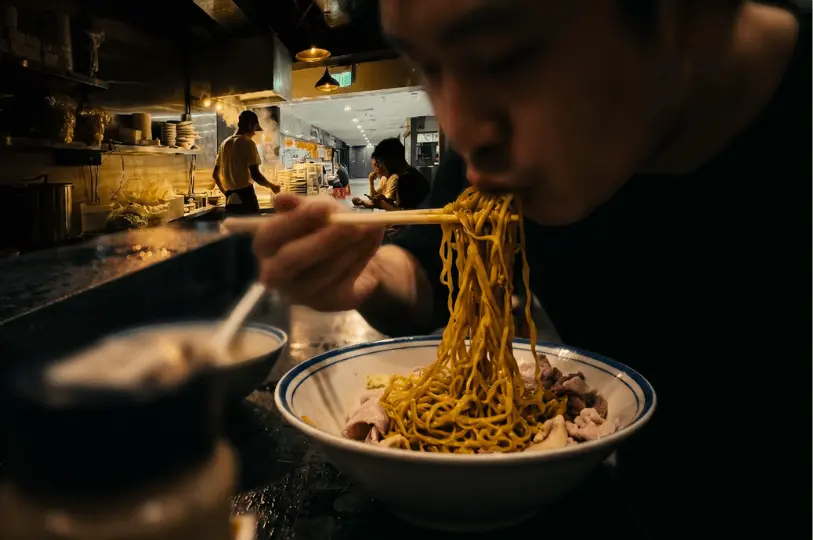 A close-up of a diner using chopsticks to lift a generous bite of yellow noodles from a bowl of Bak Chor Mee, with the steamy kitchen visible in the blurred background.