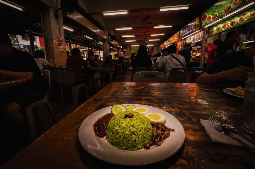 A low-angle close-up composition of green pandan Nasi Lemak rice centered on a white plate, framed by sambal, sliced hard-boiled egg, fried anchovies, and cucumber slices on a rustic wooden hawker table.