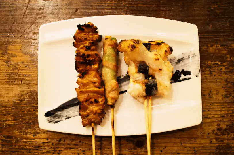 Top-down view of three assorted Japanese yakitori skewers, including grilled chicken skin and meat-wrapped vegetables, served on a white rectangular plate with a painted black brushstroke design resting on a rustic wooden table.