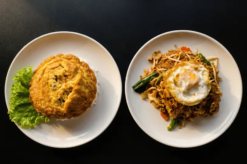 Two classic Thai dishes viewed from above on a black surface: a fluffy, golden Thai omelet over rice on the left, and a portion of stir-fried noodles topped with a fried egg on the right.