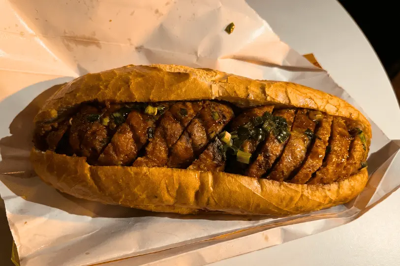 A close-up shot of a crusty bánh mì sandwich generously filled with thick, overlapping slices of glazed roasted meat and garnished with chopped scallions, resting on white wrapping paper.