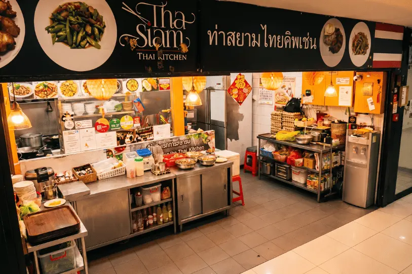 The brightly lit, open-concept kitchen and service counter of Tha Siam Thai Kitchen, showing organized stainless steel prep stations beneath the restaurant's black signboard.