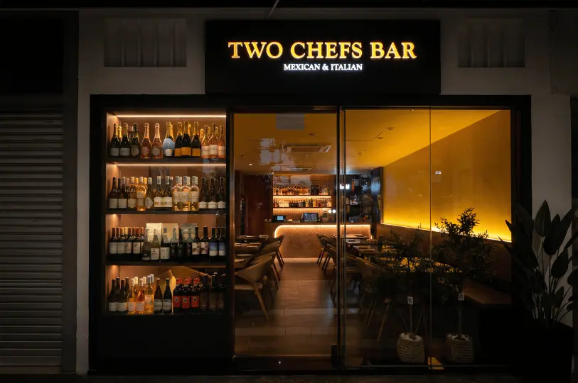 The sleek, modern glass storefront of Two Chefs Bar Mexican & Italian at night, radiating a warm, inviting yellow glow from its interior dining room and featuring a tall display shelf fully stocked with wine and liquor bottles.