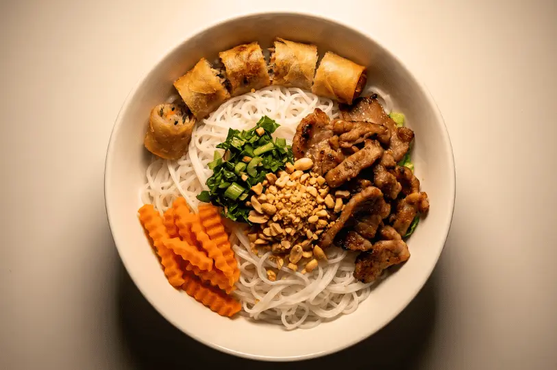 A top-down view of a white bowl containing a Vietnamese rice vermicelli dish. It is neatly arranged with grilled sliced pork, cut pieces of fried spring rolls, crinkle-cut orange carrots, fresh chopped herbs, and a mound of crushed peanuts in the center.