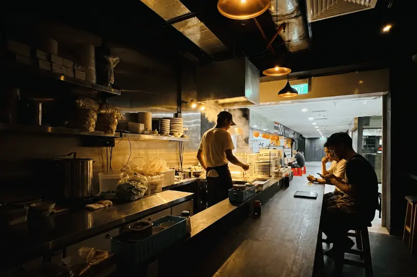 A chef cooks amidst billowing steam inside the stainless steel kitchen of Orchard Bak Chor Mee, while customers dine at the attached wooden counter.