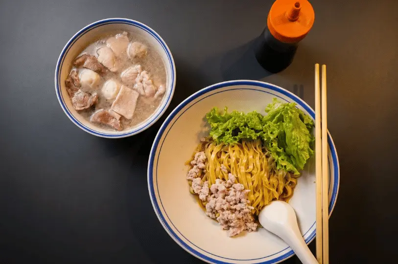 A top-down view of a Bak Chor Mee meal, featuring a bowl of flat yellow noodles topped with minced pork and lettuce, served alongside a separate bowl of rich broth filled with meatballs and sliced pork.