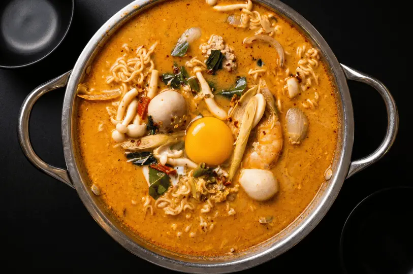 A large metal pot filled with rich, orange-colored Tom Yum noodle soup, topped with a raw egg yolk, shrimp, mushrooms, and fishballs against a dark background.