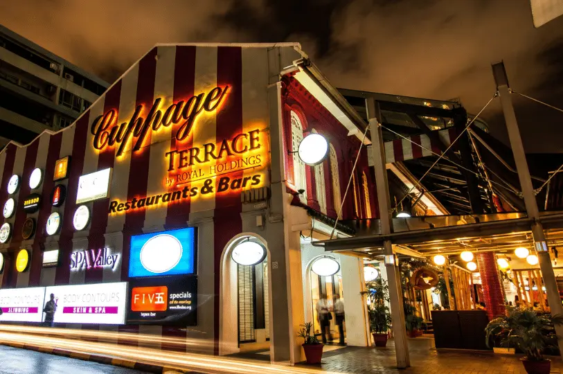 A vibrant nighttime shot of Cuppage Terrace, showcasing its distinctive red and white striped facade covered in glowing restaurant and bar signs, with long-exposure light trails from passing traffic blurring in the foreground.