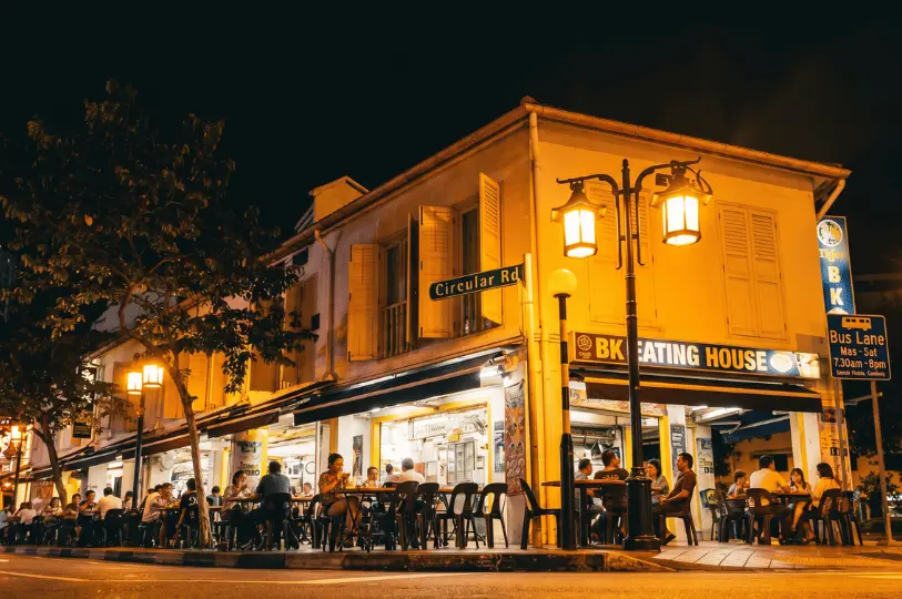 A bustling late-night scene at the brightly lit BK Eating House on Circular Road, with crowds of people dining at outdoor tables along the sidewalk.