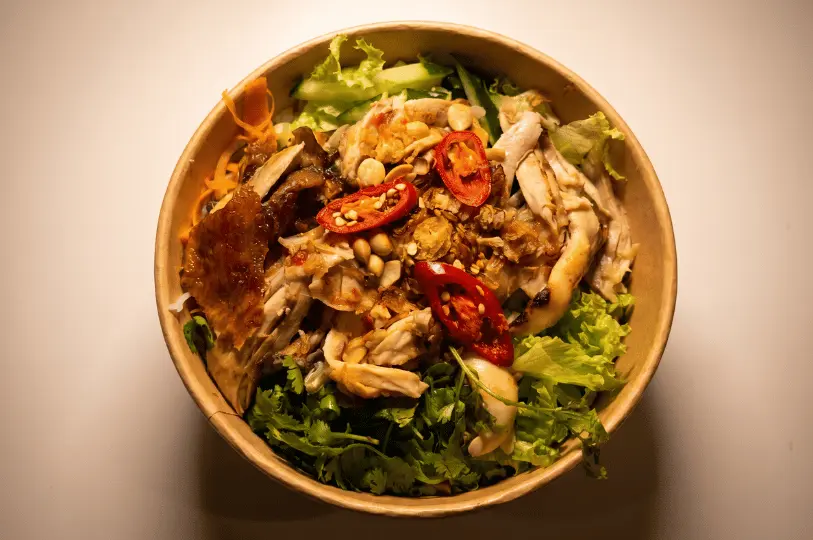 An overhead view of a vibrant Vietnamese salad or noodle bowl in a round brown paper container. It is topped with shredded chicken, crispy fried shallots, crushed peanuts, fresh cilantro, lettuce, and bright red chili slices.