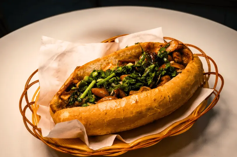 A crusty bánh mì sandwich sitting in a small woven basket lined with paper. The baguette is stuffed with a rich, dark filling of braised meats or offal and heavily garnished with fresh chopped greens.
