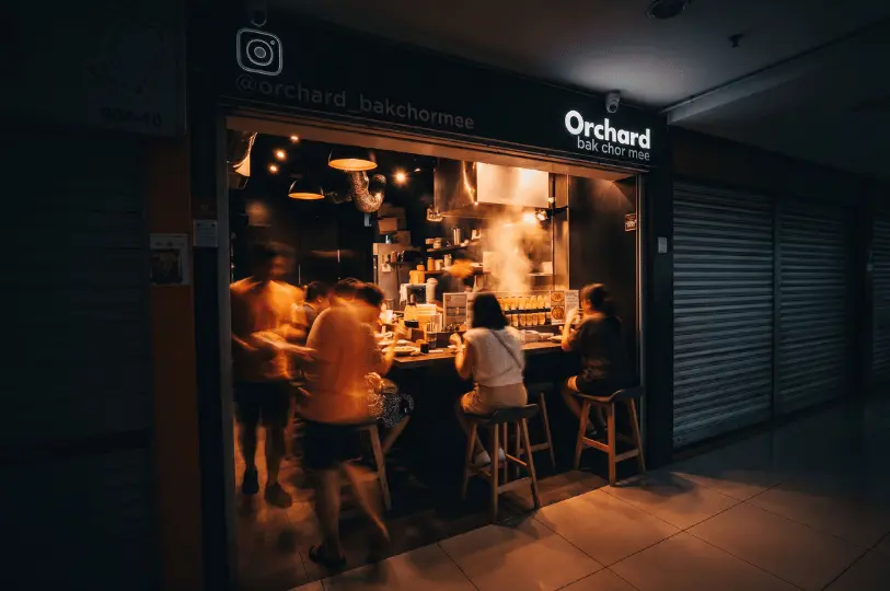 A motion-blurred view of customers crowding the glowing Orchard Bak Chor Mee stall late at night, highlighting the bustling queue nestled between closed shop shutters.