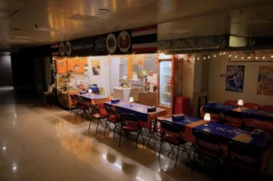 An empty, softly lit dining area of Tha Siam Thai Kitchen situated in a mall hallway, featuring long tables draped in blue and orange tablecloths beneath warm string lights.
