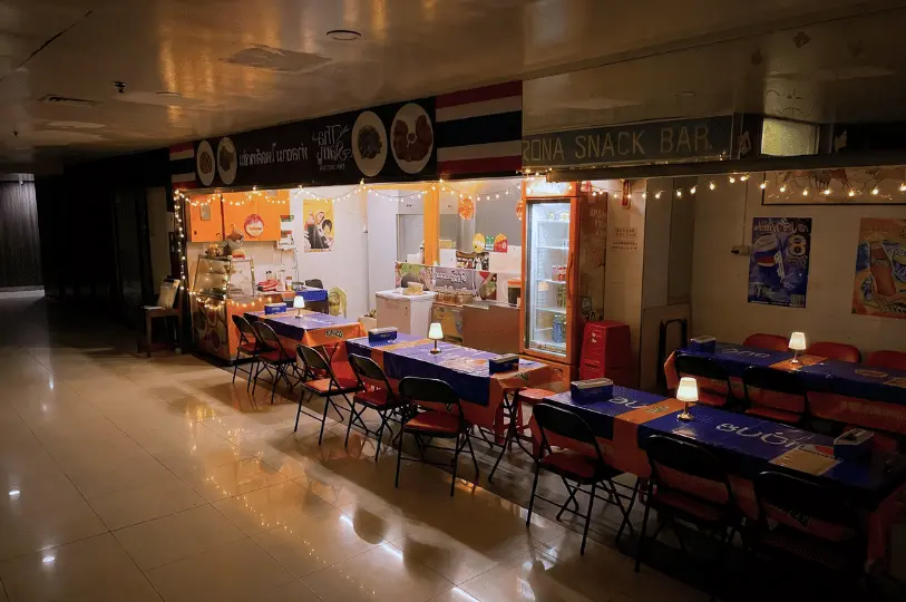An empty, softly lit dining area of Tha Siam Thai Kitchen situated in a mall hallway, featuring long tables draped in blue and orange tablecloths beneath warm string lights.