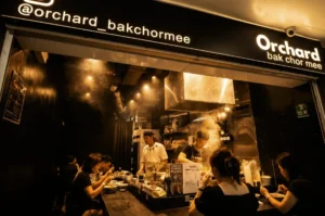 Diners sitting at a dark counter enjoying bowls of noodles right in front of the steamy, brightly lit open kitchen of the Orchard Bak Chor Mee stall.