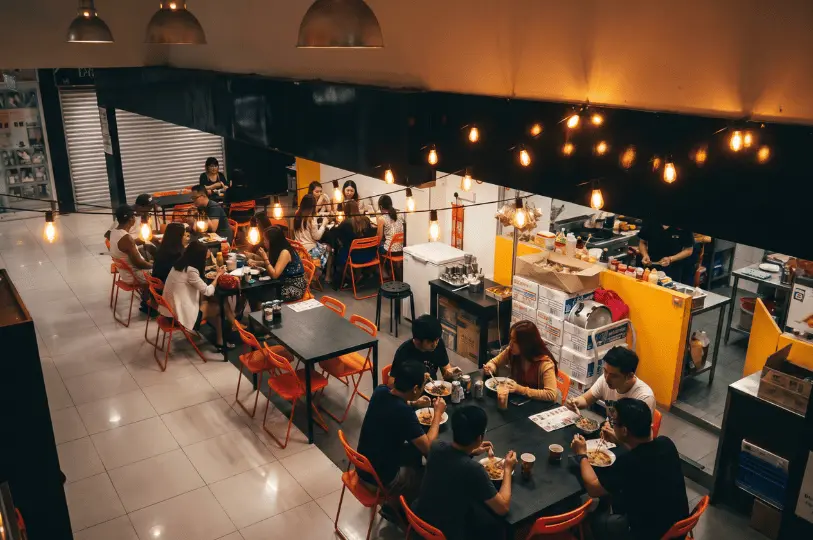 A bustling, high-angle view of diners enjoying a Thai supper at Tha Siam Thai Kitchen, illuminated by a warm glow from overhead string lights in the open concourse of Orchard Plaza.