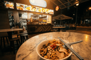 A close-up foreground shot of a steaming bowl of minced meat noodles (bak chor mee) topped with sliced pork, mushrooms, lard, and fresh red chilies on a marble table. In the background, the glowing neon sign and bright counter of an open-air Teochew Fishball Noodle stall illuminate the quiet night street.