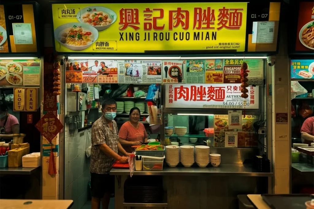 Xing Ji Ruo Cuo Mian store exterior with lighted signage and menu visible along with the vendors preparing Bak Chor Mee located at Bedok 85.