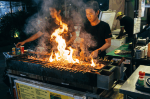 Two focused food vendors grilling rows of satay skewers over a roaring open-flame charcoal grill at night, working quietly in tandem while surrounded by billowing smoke at a bustling street food stall.