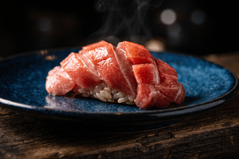 A close-up, moody shot of a single piece of glistening, deeply marbled fatty tuna (otoro) nigiri resting on a mottled blue ceramic plate over rustic wood, with faint steam rising into the dark background.