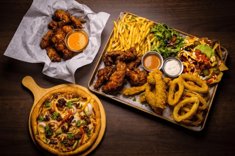 A sprawling, late-night bar food spread arranged on a dark wooden table. The high-contrast image features a massive sharing tray loaded with crispy fried chicken wings, fries, calamari rings, chicken tenders, and fully loaded nachos, accompanied by a rustic meat pizza and extra wings in a basket.