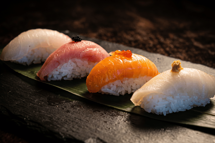 Four distinct pieces of nigiri—including tuna, salmon topped with roe, and white fish—arranged diagonally on a vibrant green leaf over a textured dark stone surface under dramatic spotlighting.