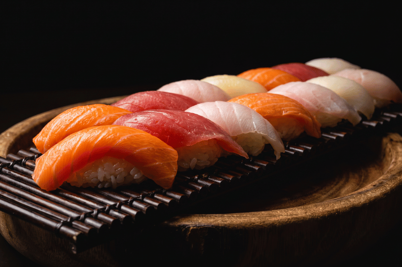 A tightly packed row of assorted nigiri sushi, featuring vibrant slices of salmon, tuna, and white fish, presented on a slatted bamboo mat within a carved wooden bowl against a stark black background.