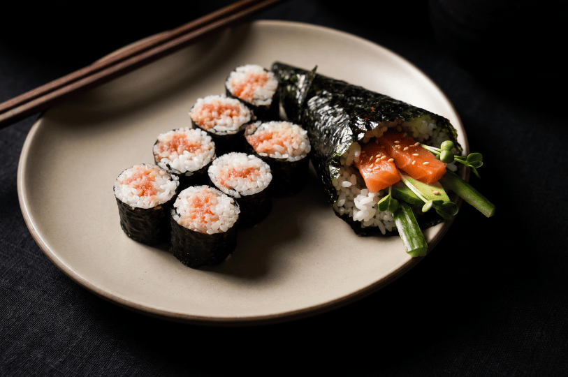 A minimalist pale ceramic plate against a dark setting, holding six pieces of chopped tuna maki alongside a large, cone-shaped hand roll filled with salmon, cucumber, and fresh greens.