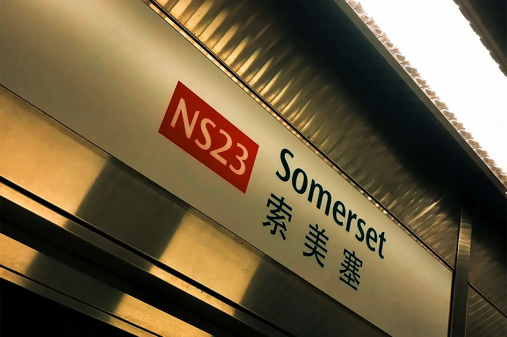 Somerset MRT Station entrance on Orchard Road at night, with illuminated signs and pedestrians passing through the surrounding shopping district.
