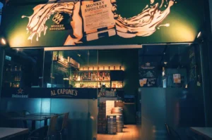 The dimly lit, urban exterior of Al Capone's bar at Cuppage Terrace. Warm indoor light spills through the glass storefront, illuminating stacked metal kegs and empty outdoor dining tables, while a large, vibrant Monkey Shoulder whisky mural dominates the space above the entrance.