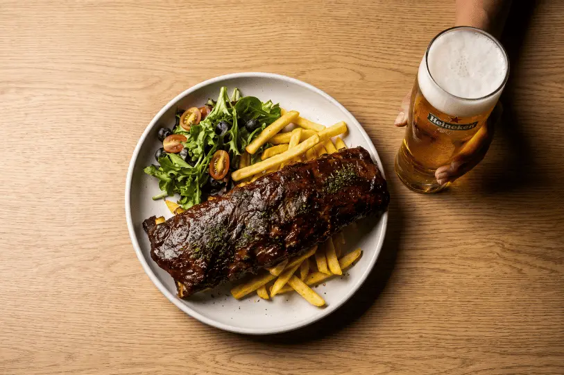 A top-down view on a light oak table featuring a plate of sticky, dark-glazed barbecue ribs resting on golden French fries, paired with a fresh side salad. On the right edge of the frame, a hand holds a chilled, frothy pint of Heineken beer, capturing a casual dining moment.
