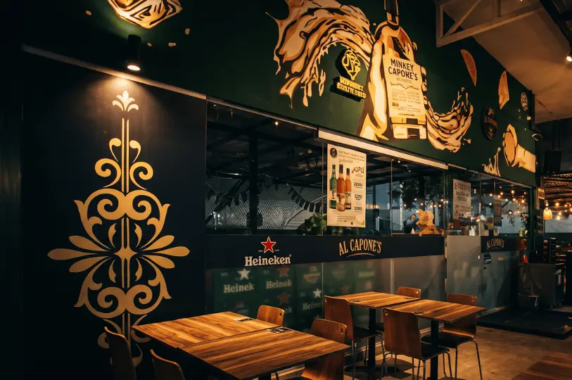 The atmospheric outdoor seating area of Al Capone's at night. Warm spotlights cast a glow over empty wooden tables and chairs, contrasting against the dark walls adorned with an intricate gold stencil design and a lively, painted liquor mural stretching across the upper facade.