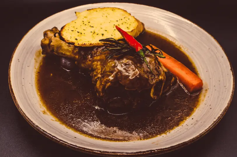 A close-up, moody food photograph highlighting the rich textures of a slow-braised bone-in meat dish submerged in a glossy, dark gravy. The rustic plate is garnished with toasted garlic bread, a whole carrot, fresh rosemary, and a vibrant red chili to contrast the deep, warm tones.