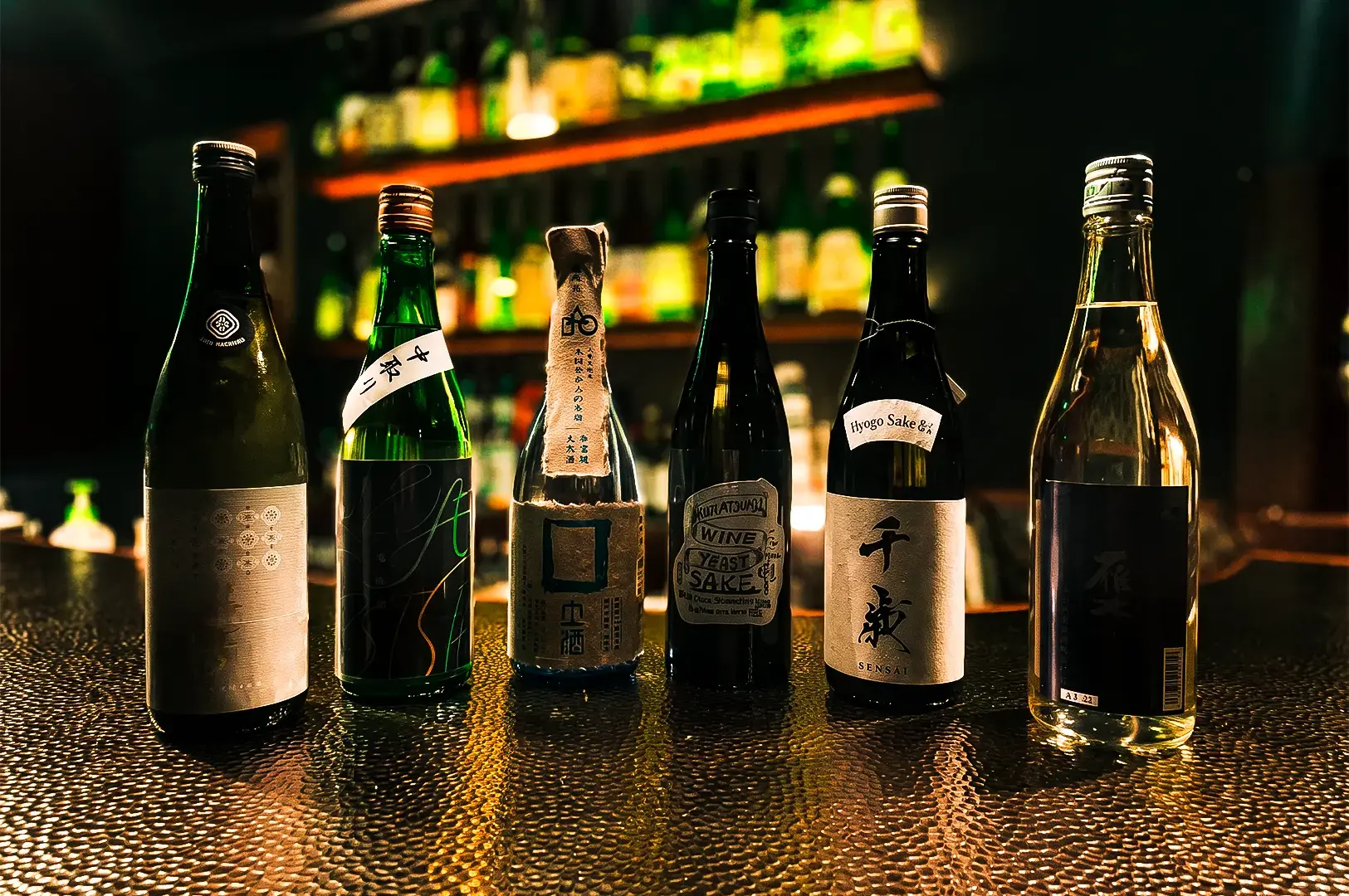 A variety of sake bottles lined up on a table illuminated by dramatic dim lights with other bottles placed on a shelf visible in the background.
