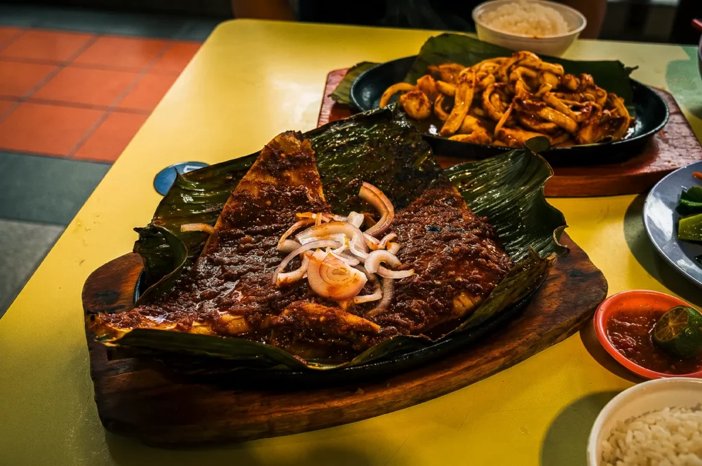 Chomp Chomp BBQ Signature Sambal BBQ Stingray plated on a banana leaf and wooden platter together with side dishes and sauces on a yellow table at Chomp Chomp BBQ. Bedok 85, Singapore.