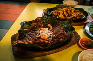 Chomp Chomp BBQ Signature Sambal BBQ Stingray plated on a banana leaf and wooden platter together with side dishes and sauces on a yellow table at Chomp Chomp BBQ. Bedok 85, Singapore.