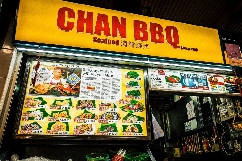 Chan BBQ exterior focusing on the shop’s lighted logo signed in yellow in red with its lighted menu board below showing menu photos and prices.