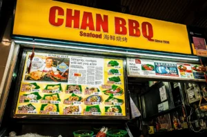 Chan BBQ exterior focusing on the shop’s lighted logo signed in yellow in red with its lighted menu board below showing menu photos and prices.